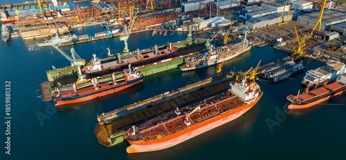 Side view drone shot of a large orange tanker ship lifted in a floating dry dock for hull inspection and painting surrounded by other docked industrial vessels