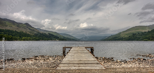A panoramic view looking down a small jetty across Loch Earn in Scotland towards the distant hills