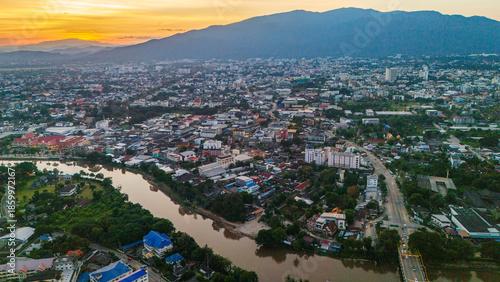 Schilderij op canvas Cinematic aerial view of Chiang Mai city at sunset, featuring the Ping River winding through the urban landscape with misty mountains in the background