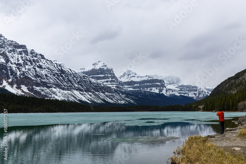 Landscape photograph of icy water in frozen river or lake at base of mountains on cloudy stormy cold day with snow coming and tourist from behind taking picture of the beautiful blue water scenery