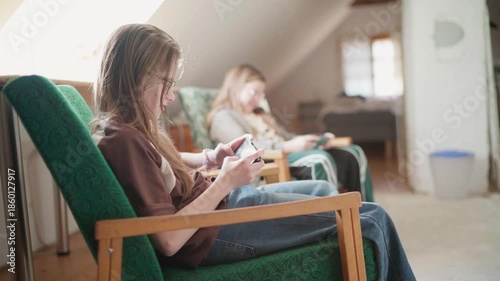 Two sisters sitting in comfortable chair in kids' room cheerfully smiling and laughing while playing online game together using smartphones connected to internet Happy children, game addiction concept