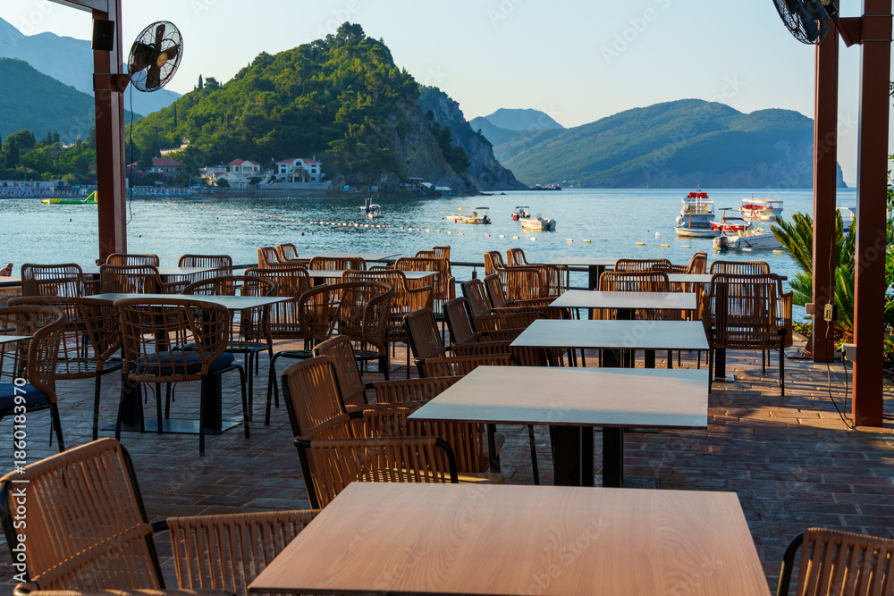 Fototapeta premium cozy outdoor cafe or restaurant located on embankment with panoramic view of Petrovac city, Montenegro, tables and chairs in front of sea and beach, beautiful nature with hills and forest