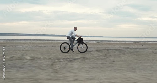 Determined businessman pedals rapidly along glowing shoreline creating energetic rhythm. Office-styled man pushes bicycle forward embracing windy evening motion