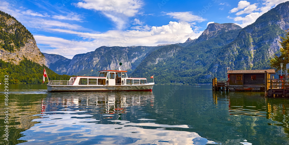 Fototapeta premium Hallstatt, Austria. Touristic pleasure boat arriving to the old wooden dock at lake Hallstattersee among austrian Alps mountains. Picturesque summery landscape. Blue sky with clouds.