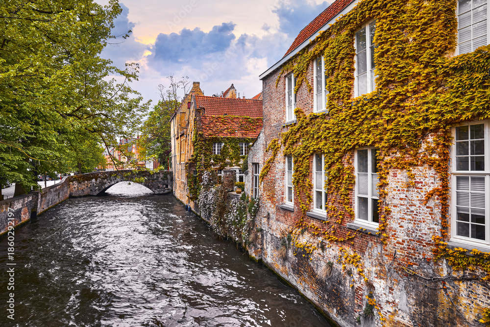 Fototapeta premium Bruges, Belgium. Medieval vintage brick houses with balconies over river with old stone bridge, covered with green plants and bushes. Summer landscape with sky and clouds..