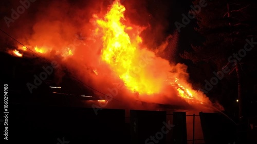 A house fire erupts at night, engulfing the structure in flames and smoke, creating a dramatic and dangerous situation for nearby residents.