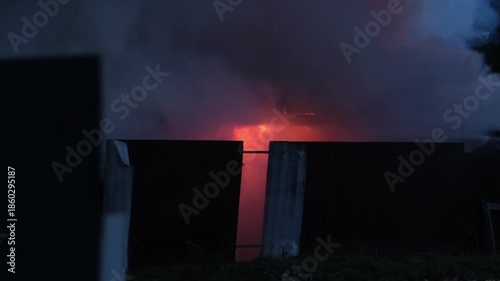 A large house fire erupts at night, with flames and thick smoke affecting the nearby homes and the communitys safety.