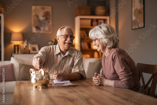 Tableau sur toile Warm Home Moment: Elderly Couple Smiles At Table With Piggy Bank And Papers