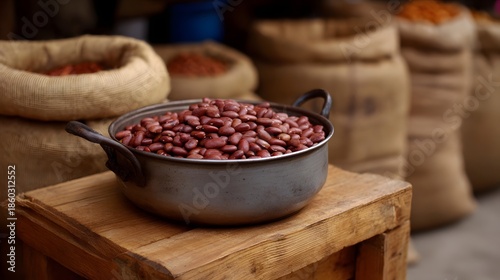 A close up view of a pot filled with raw red beans resting on a wooden crate with blurred sacks of grain in the background