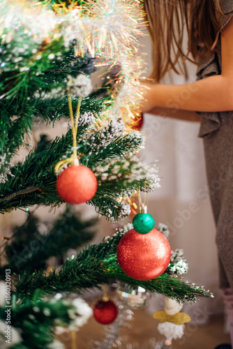 A child hangs a shiny Christmas ball, an ornament hanging from dark green branches, creates a festive atmosphere.