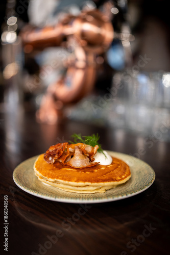 Stack of golden American-style pancakes with crispy bacon pieces, a dollop of white cream or crème fraîche, and a sprig of fresh dill on a patterned plate in a moody cafe setting.