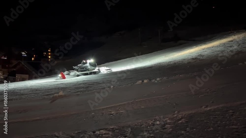 A brightly lit snowcat moves up the ski slope, compacting the snow beneath it