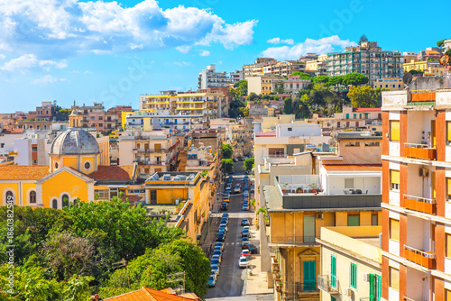 Densely packed cityscape on steep hill, likely in Italian city of Messina, Sicily. Scene is filled with traditional Mediterranean buildings, narrow streets, and greenery under blue sky