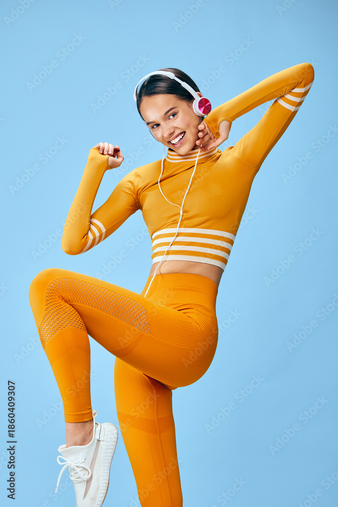 Obraz premium Fitness woman in vibrant orange athletic outfit performs dynamic pose against blue backdrop. Sporty wear, headphones, and energetic movement highlight cardio training, dance-inspired workout,