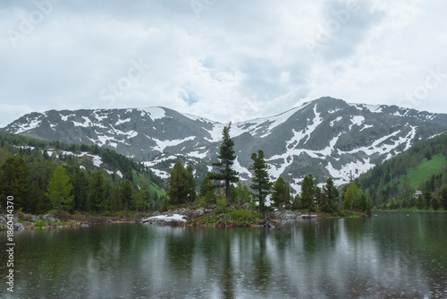 Alpine lake and coniferous forest against high rocky snowy mountain wall unde...