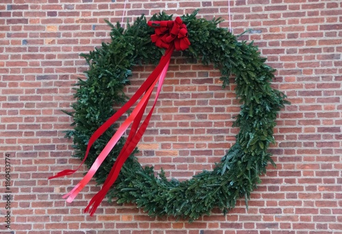 A festive pine wreath with cascading red and pink ribbons, displayed on brick wall to celebrate the holiday season.