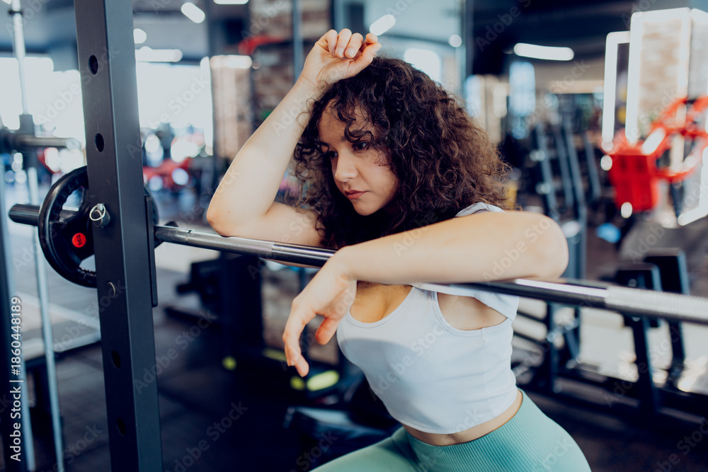 Fototapeta premium Tired female athlete taking a break, resting her head on a weight bar after intense strength training