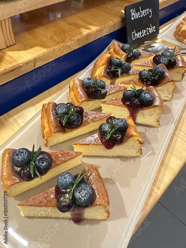 Close up of sliced blueberry cheesecake arranged in a row with fresh blueberries and sauce drizzle, showing creamy texture and warm bakery display atmosphere.