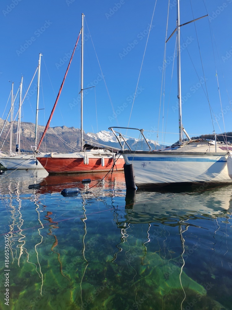Fototapeta premium Sailing boats and yachts are at the harbour of Annecy lake. Snow-capped mountains Mont Baret in the background