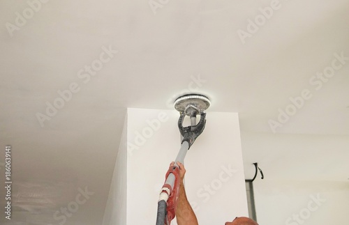 Sanding a ceiling during a home renovation.A construction worker sands a ceiling with an electric drywall sander. Electric drywall sander with LED light.