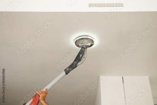 Sanding a ceiling during a home renovation.A construction worker sands a ceiling with an electric drywall sander. Electric drywall sander with LED light.
