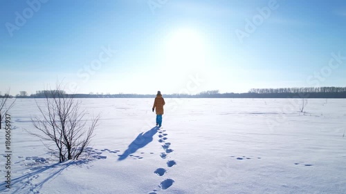 Rear view of a woman walking alone across a vast snowy field, leaving footprints on fresh snow. Minimal winter landscape with clear sky, solitude, freedom, journey and calm concept.