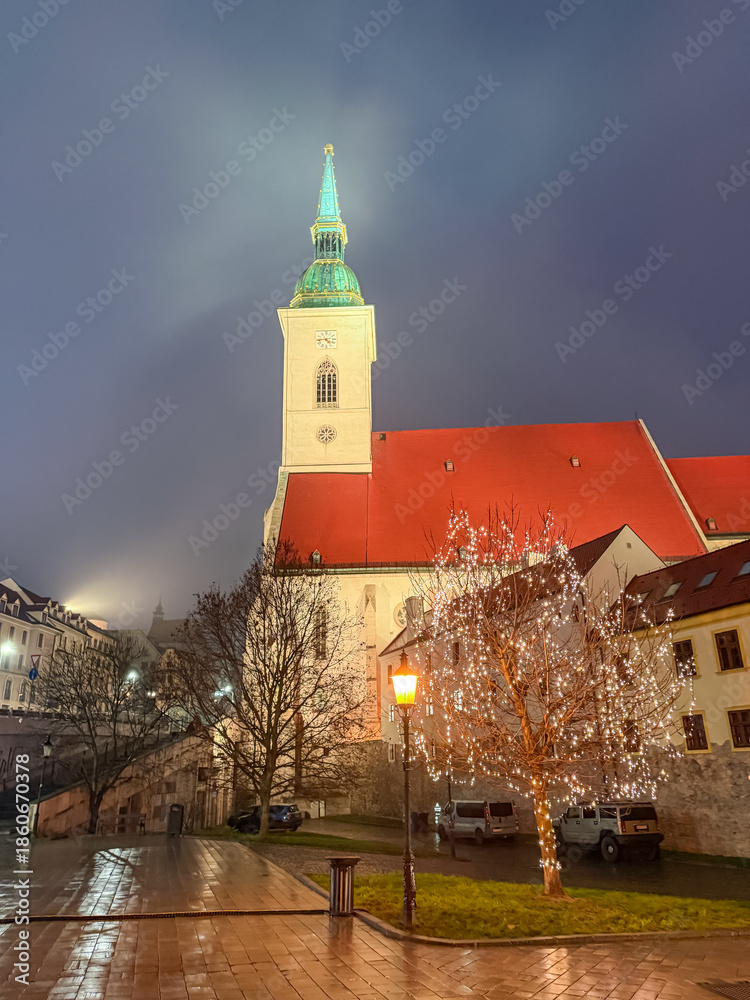 Obraz premium Illuminated St. Martin's Cathedral tower rising above a glowing tree in Bratislava, Slovakia, casting reflections on wet cobblestones during a foggy Christmas night in the historic city center.