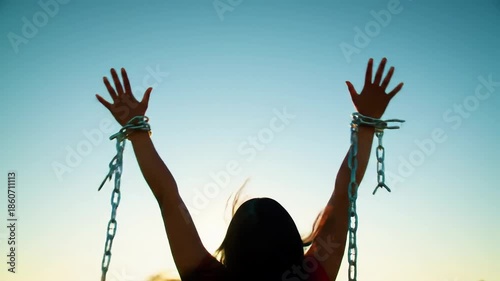 Woman in silhouette with arms raised, breaking metal chains around her wrists against a clear sky, symbolizing liberation and freedom journey