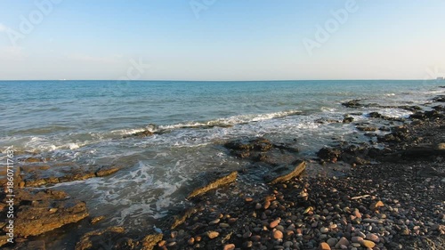 Rocky sea shoreline with small waves washing over stones, calm coastal landscape under clear blue sky, natural seascape, no people, sunny weather, daytime outdoor scene
