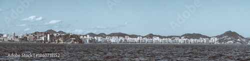 Telephoto panoramic view of Niteroi waterfront skyline across Guanabara Bay, modern apartment towers and green hills under blue sky, textured sea foreground, copy space