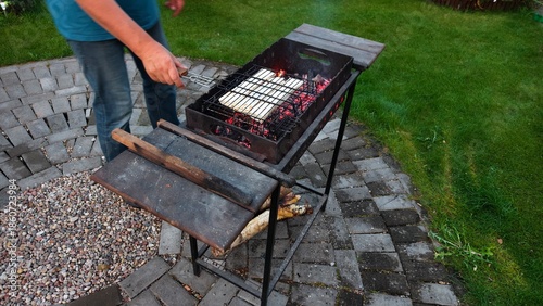 On a tiled area near a grassy lawn there is a metal brazier in which coals are smoldering and the light burns. A man flips a grate of cheese in a pita bread. Autumn evening twilight
