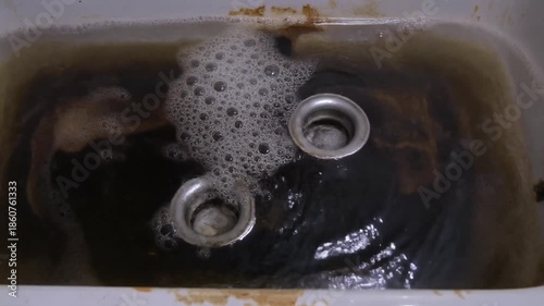 A close-up view of a dirty sink filled with murky brown water, showing submerged dark items and foam on the surface.