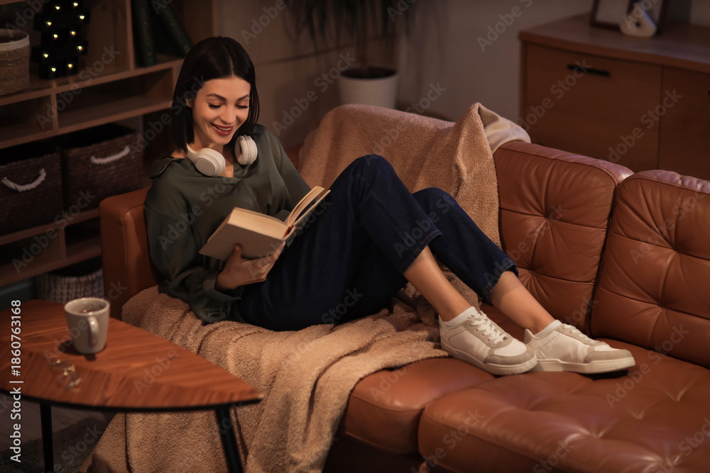 Obraz premium Beautiful young woman with headphones reading book and sitting on sofa in living room at night