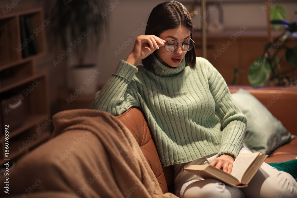 Fototapeta premium Beautiful young woman in eyeglasses reading book and sitting on sofa in living room at night