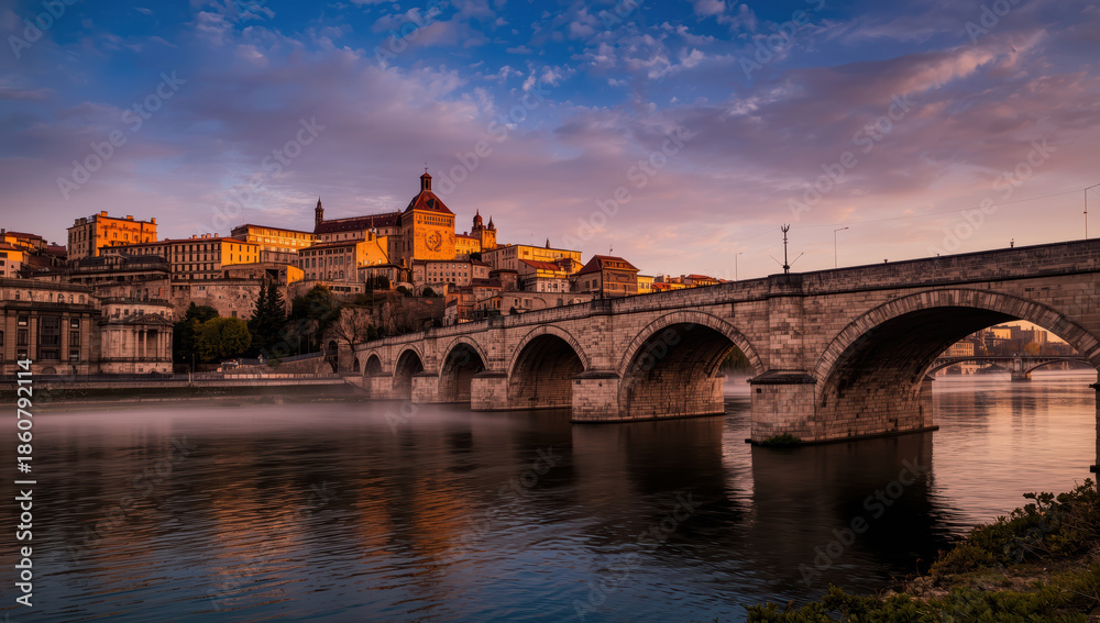 Obraz premium Historic city skyline at golden hour with arched stone bridge and reflective river