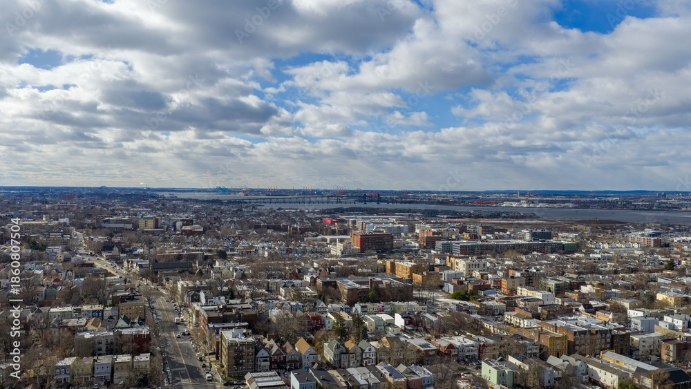 Fototapeta premium Aerial drone photo from Jersey City featuring Manhattan skyline views, dense city blocks, waterfront buildings, and modern New York City architecture under dynamic daytime cloud cover. 