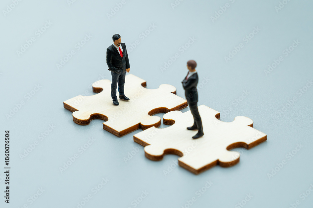 Fototapeta premium Miniature business figures stand on interlocking puzzle pieces. Corporate partnership, teamwork, strategic collaboration concept.