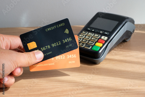 Hand holding yellow and black credit cards in front of an electronic EDC terminal on a wooden desk. Perfect for fintech, payment systems, ecommerce, business finance, banking services, technology