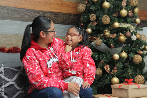 Latina single mom with her 4-year-old brunette daughter wearing glasses, dressed in ugly sweaters, open presents as a family next to the Christmas tree