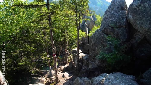A narrow, rocky hiking trail winds through dense forest and large boulders, ascending towards the distant peaks of Mt. Katahdin in Maine on a bright, sunny day.
