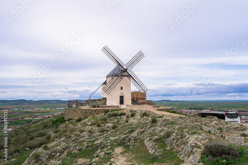Traditional white windmill with restored sails perched on a rocky hill along the famous Don Quixote Route, overlooking green agricultural plains in spring.