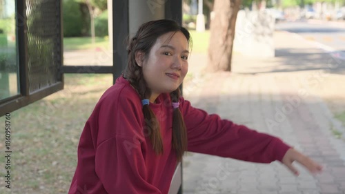 Asian woman smiling and raising hand to call car taxi or friend at bus stop during daytime urban lifestyle