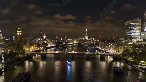 Aerial night view of a cityscape along a river, with illuminated buildings, bridges, and boats reflecting in the water under a cloudy sky