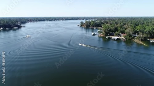 Aerial shot overlooking a tranquil lake with boats creating wakes. Lush green trees line the shore. Clear sky and calm water