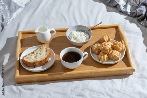Morning meal on a wooden tray with bread, coffee, cream, and pastries on a bed