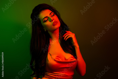 Glamorous young woman with dark hair in satin blouse illuminated by neon green and red light in artistic background