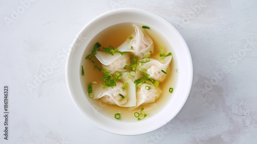 Minimalist overhead shot of Chinese wonton soup. Delicate shrimp and pork dumplings in a clear, golden broth garnished with fresh green onions in a white bowl.