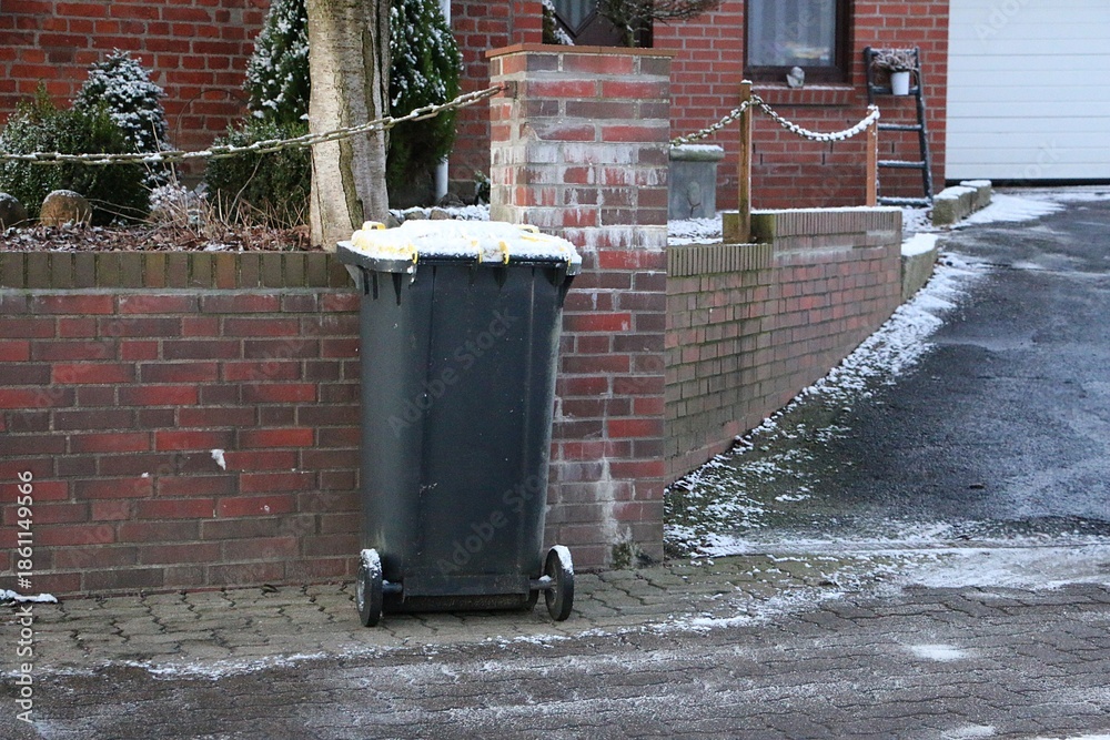Obraz premium A garbage can, covered with a little snow, stands against the wall on the sidewalk and is ready for collection