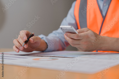 Принти на полотні Engineer or construction worker wearing safety vest, holding a smartphone and pen while reviewing documents on a desk