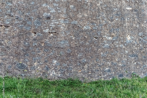 Textured stone wall with embedded rocks meets vibrant green grass, creating strong contrast and ample copy space. An ideal background for design elements, text overlay, or product placement